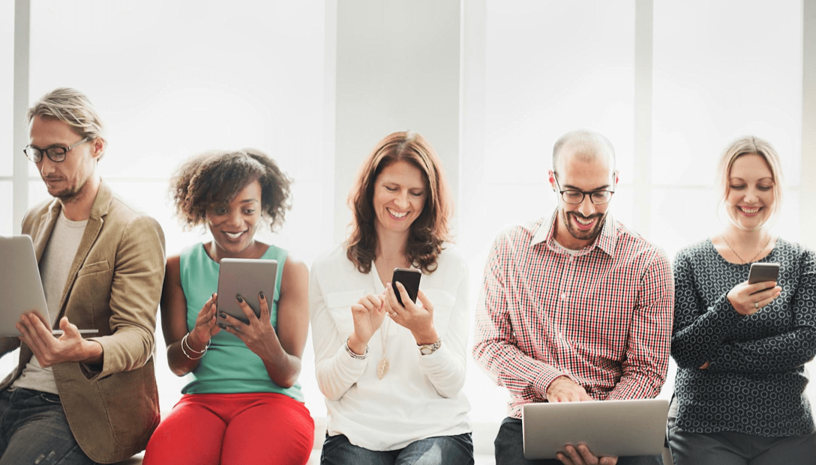 Five people sitting in a bright coworking space, each using a laptop, tablet, or smartphone.